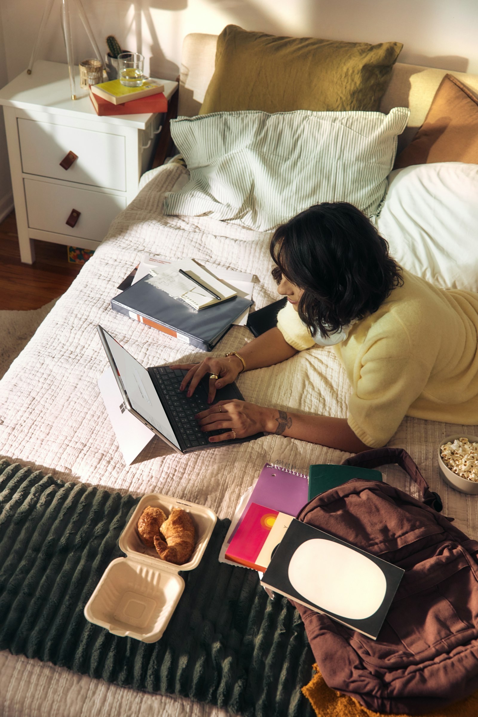Woman working on a laptop in a relaxed home setting with books and snacks nearby.
