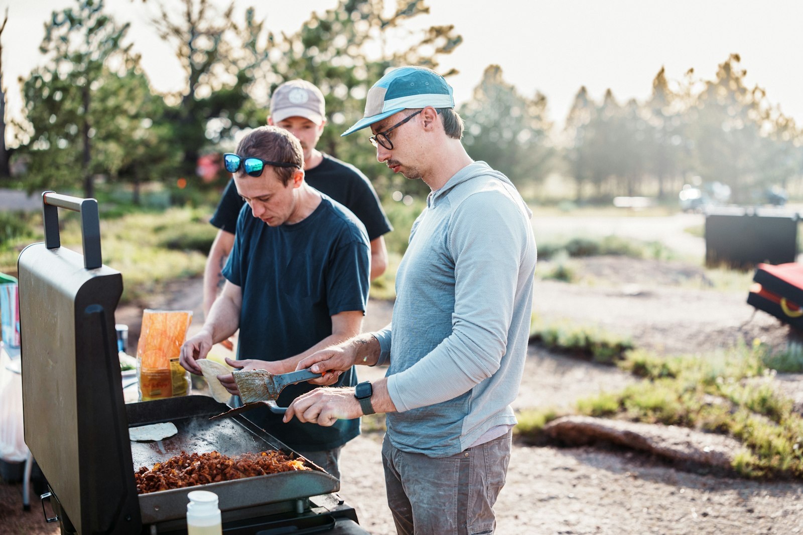 Three people cooking together outdoors around a grill.